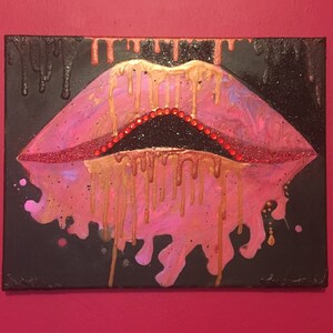 May include: A rectangular canvas painting featuring a pair of stylized lips in shades of pink and red. The lips are accented with gold paint drips and small red rhinestones along the inner edge. The background is black with gold paint drips.