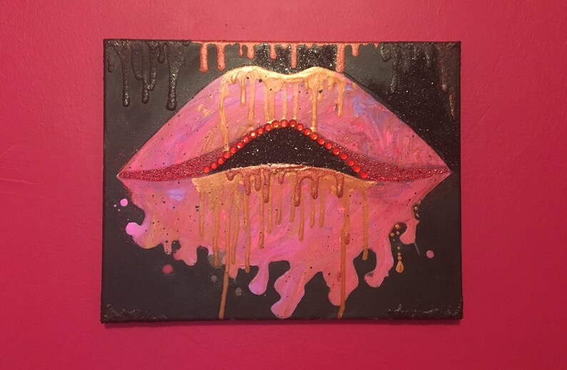 May include: A rectangular canvas painting featuring a pair of stylized lips in shades of pink and red. The lips are accented with gold paint drips and small red rhinestones along the inner edge. The background is black with gold paint drips.
