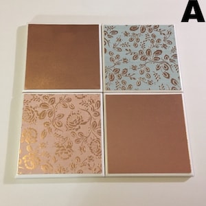 Puede incluir: Conjunto de cuatro posavasos cuadrados de cer&aacute;mica con borde blanco. Dos posavasos tienen un acabado met&aacute;lico cobre y dos posavasos tienen un patr&oacute;n floral con detalles dorados. Un posavasos tiene un fondo azul claro con un patr&oacute;n floral dorado, y el otro tiene un fondo rosa con un patr&oacute;n floral dorado.