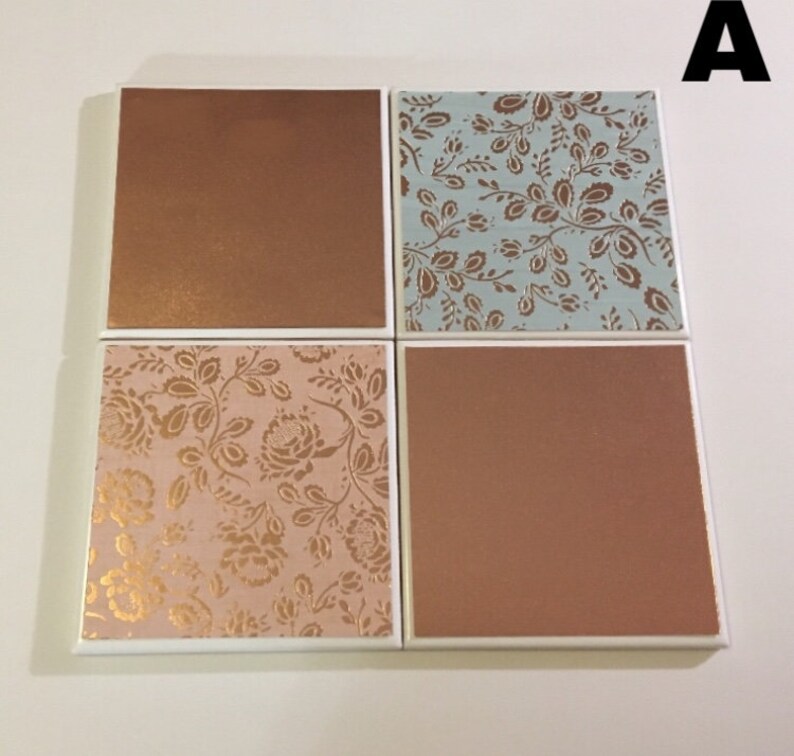 Puede incluir: Conjunto de cuatro posavasos cuadrados de cer&aacute;mica con borde blanco. Dos posavasos tienen un acabado met&aacute;lico cobre y dos posavasos tienen un patr&oacute;n floral con detalles dorados. Un posavasos tiene un fondo azul claro con un patr&oacute;n floral dorado, y el otro tiene un fondo rosa con un patr&oacute;n floral dorado.
