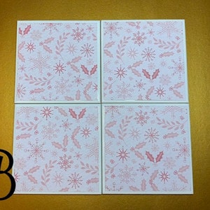 Peut inclure: Quatre sous-verres carr&eacute;s blancs avec un motif de flocons de neige et de feuilles de houx roses et blancs.