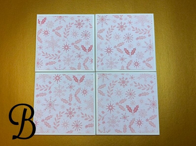 Peut inclure: Quatre sous-verres carr&eacute;s blancs avec un motif de flocons de neige et de feuilles de houx roses et blancs.