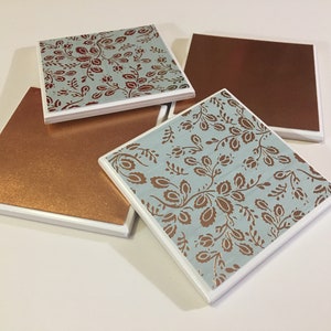Puede incluir: Cuatro posavasos cuadrados de cer&aacute;mica con borde blanco. Dos posavasos tienen un fondo azul con un patr&oacute;n floral de color cobre. Los otros dos posavasos son de cobre s&oacute;lido.