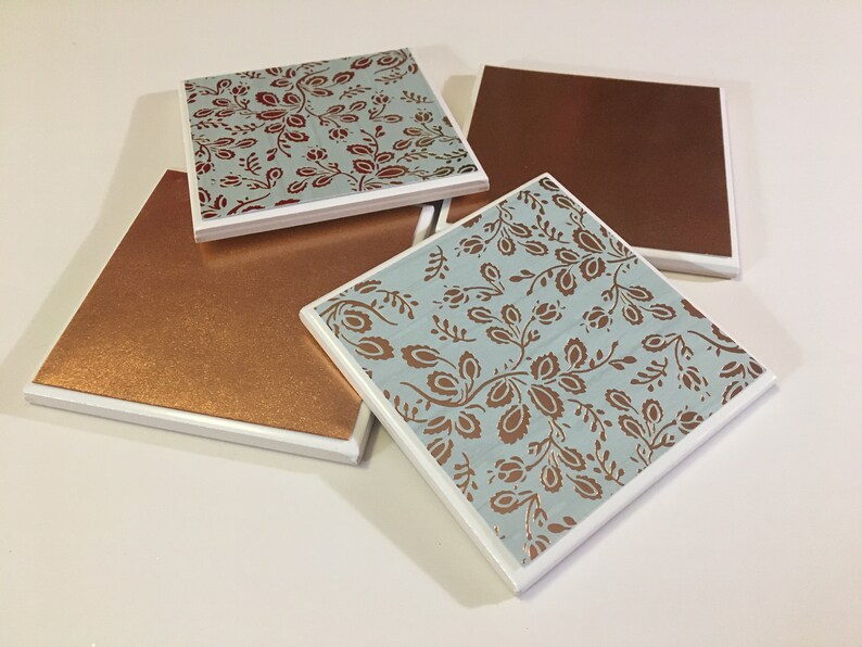 Puede incluir: Cuatro posavasos cuadrados de cer&aacute;mica con borde blanco. Dos posavasos tienen un fondo azul con un patr&oacute;n floral de color cobre. Los otros dos posavasos son de cobre s&oacute;lido.