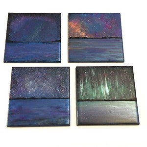 Puede incluir: Conjunto de cuatro posavasos cuadrados con im&aacute;genes pintadas de noches estrelladas y agua. Cada posavasos presenta una escena de cielo nocturno diferente con un reflejo de agua azul oscuro.