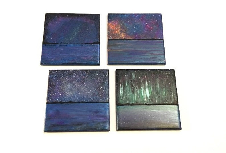 Puede incluir: Conjunto de cuatro posavasos cuadrados con im&aacute;genes pintadas de noches estrelladas y agua. Cada posavasos presenta una escena de cielo nocturno diferente con un reflejo de agua azul oscuro.
