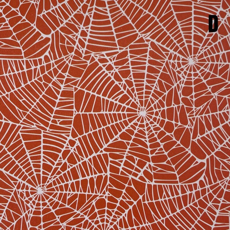 K&ouml;nnte beinhalten: Eine flache Stoffoberfl&auml;che mit einem sich wiederholenden Muster aus wei&szlig;en Spinnennetzen auf einem einfarbigen, gebrannten orangefarbenen Hintergrund. Das Design umfasst mehrere &uuml;berlappende Netzstrukturen und der Buchstabe D ist in der oberen rechten Ecke aufgedruckt.