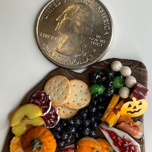 Op de afbeelding: Een miniatuur Halloween-charcuteriebord met een verscheidenheid aan lekkernijen, waaronder crackers, kaas, fruit, salami en snacks in de vorm van pompoenen. Een Amerikaanse kwartje is opgenomen ter vergelijking.