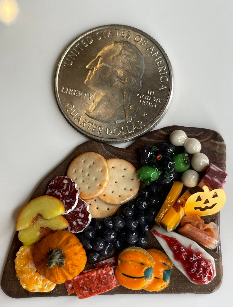 Op de afbeelding: Een miniatuur Halloween-charcuteriebord met een verscheidenheid aan lekkernijen, waaronder crackers, kaas, fruit, salami en snacks in de vorm van pompoenen. Een Amerikaanse kwartje is opgenomen ter vergelijking.