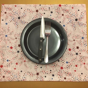 Peut inclure: Un set de table blanc avec un motif d'&eacute;toiles rouge, blanc et bleu. Une assiette noire avec une fourchette et un couteau argent&eacute;s est pos&eacute;e sur le set de table.