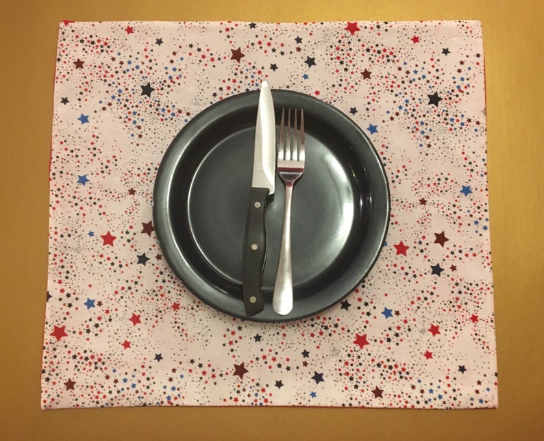 Peut inclure: Un set de table blanc avec un motif d'&eacute;toiles rouge, blanc et bleu. Une assiette noire avec une fourchette et un couteau argent&eacute;s est pos&eacute;e sur le set de table.
