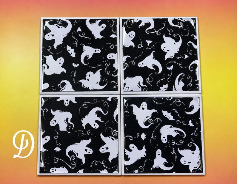 May include: A set of four square coasters featuring a repeating pattern of white ghosts and small bats on a black background. Each coaster has a glossy finish and a white border. The letter D is visible in white on the bottom left corner.