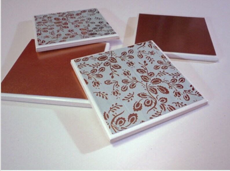 Puede incluir: Cuatro posavasos cuadrados de cer&aacute;mica con borde blanco. Dos posavasos presentan un patr&oacute;n floral marr&oacute;n sobre un fondo azul claro. Dos posavasos son de color marr&oacute;n s&oacute;lido.