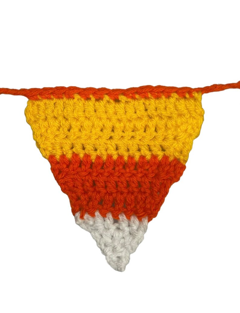 May include: A handmade crochet candy corn banner. The banner features a triangle shape with orange, yellow, and white sections, resembling candy corn. The banner is made of yarn and is suitable for Halloween or fall decor.