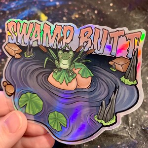 May include: Holographic sticker featuring a cartoon frog sitting on a lily pad in a pond. The sticker has the words "SWAMP BUTT" in a colorful, bubble-style font. The frog is green, and the pond is a mix of blues and purples.