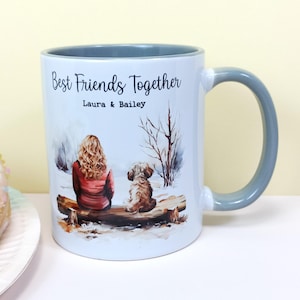 May include: White ceramic mug with a grey handle. The mug features a design of a woman and a dog sitting on a bench in a snowy woodland. The text "Best Friends Together" is printed on the mug, along with the names "Laura & Bailey".