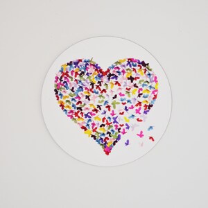 May include: A heart-shaped design composed of colorful butterfly illustrations on a white circular background. The butterflies are in shades of pink, blue, yellow, and purple. Some butterflies appear to be flying away from the heart.