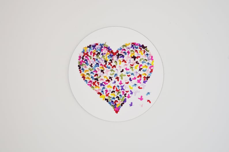 May include: A heart-shaped design composed of colorful butterfly illustrations on a white circular background. The butterflies are in shades of pink, blue, yellow, and purple. Some butterflies appear to be flying away from the heart.