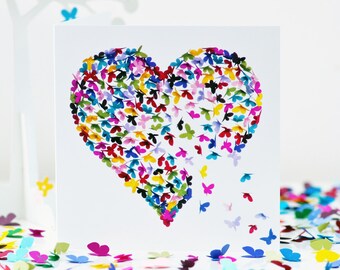 May include: A white card features a heart design composed of colorful paper butterflies. The butterflies are in shades of red, pink, yellow, blue, and green. Some butterflies appear to be falling away from the heart, with more scattered below.