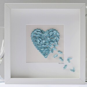 May include: A framed artwork featuring a heart shape composed of numerous light blue paper butterflies. Some butterflies appear to be fluttering away from the heart. The artwork is set against a white background within a white frame.