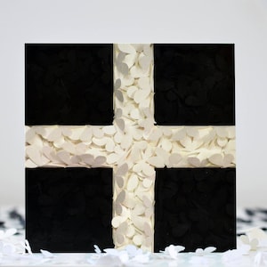 May include: A square, abstract art piece featuring a cross design. The cross is composed of white, petal-shaped elements, set against a background of four black squares. The artwork is on a white surface.