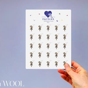 May include: A white rectangular card featuring multiple illustrations of a girl in a leopard print dress, holding a wand. The card has a blue heart logo with the text "INKYWOOL" and "The Butterfly Art Store". The card is held by a hand.