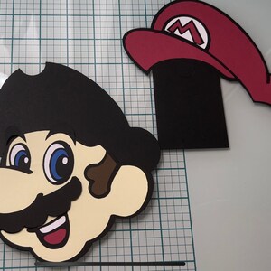 May include: A black and white papercraft of a cartoon character with a black hat and a moustache. The character has large eyes and a wide smile. The character is wearing a red hat with a white 'M' on it.