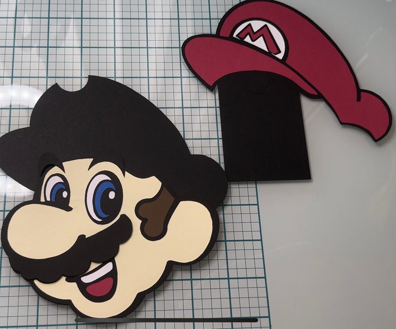 May include: A black and white papercraft of a cartoon character with a black hat and a moustache. The character has large eyes and a wide smile. The character is wearing a red hat with a white 'M' on it.