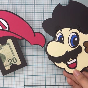 May include: Two papercraft Mario character money holders. One is a red hat with a black M on the front and the other is a brown-haired Mario with a black moustache and a red mouth. Both have a slot for a bill to be inserted.