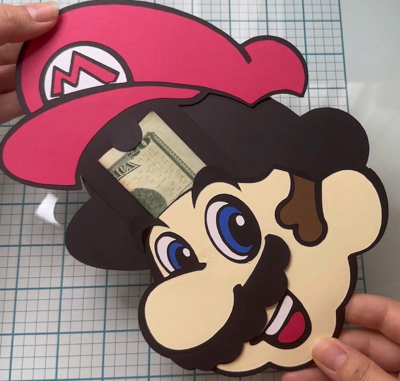 May include: A papercraft Mario head with a red hat and a dollar bill tucked inside. The head is brown, yellow, and black with blue eyes and a brown moustache.
