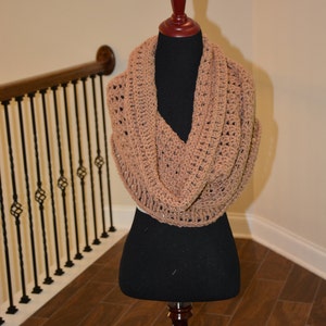 Puede incluir: Una bufanda infinita de crochet marrón, expuesta en un maniquí negro. La bufanda está hecha con un patrón de punto simple y tiene una sensación suelta y fluida.