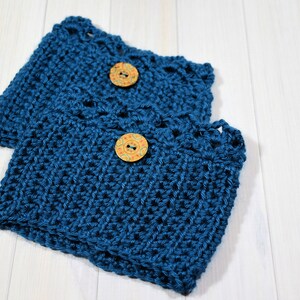May include: Two teal blue crocheted coffee cup cozies with decorative buttons. The cozies are made with a textured stitch and have a scalloped edge.