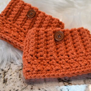 May include: Two crocheted orange leg warmers with brown buttons. The leg warmers are made with a textured stitch and are lying on a white fluffy surface.