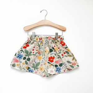 May include: Floral print shorts with a gathered elastic waistband. The shorts are beige with a variety of colorful flowers, including red, pink, blue, yellow, and green.