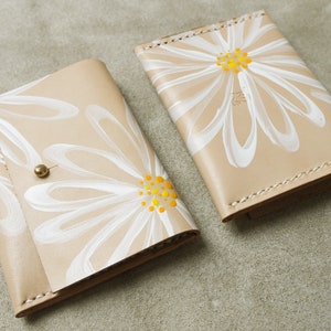 May include: Two beige leather wallets with white daisy designs. The daisies have yellow centers. One wallet has a single daisy design, while the other has multiple daisies.
