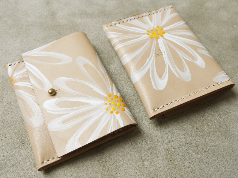 May include: Two beige leather wallets with white daisy designs. The daisies have yellow centers. One wallet has a single daisy design, while the other has multiple daisies.