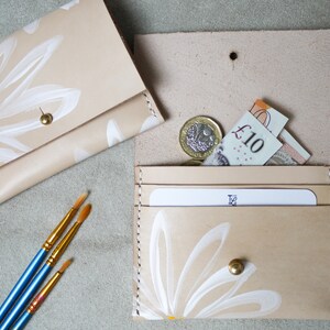 May include: Two beige leather wallets with white daisy designs. The wallet on the left is closed and the wallet on the right is open, showing a card slot and a pocket for cash. The wallet on the right also has a gold button.