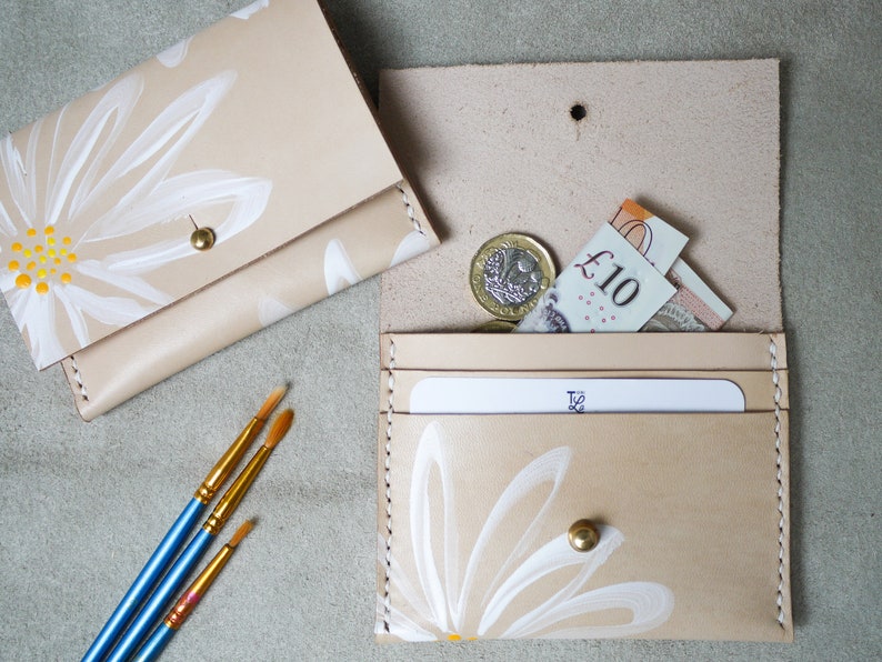 May include: Two beige leather wallets with white daisy designs. The wallet on the left is closed and the wallet on the right is open, showing a card slot and a pocket for cash. The wallet on the right also has a gold button.