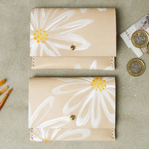 May include: Two beige leather wallets with white daisy designs and yellow centers. The wallets have a gold snap closure.