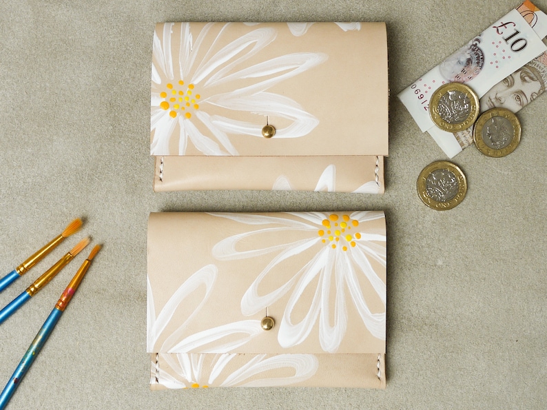 May include: Two beige leather wallets with white daisy designs and yellow centers. The wallets have a gold snap closure.