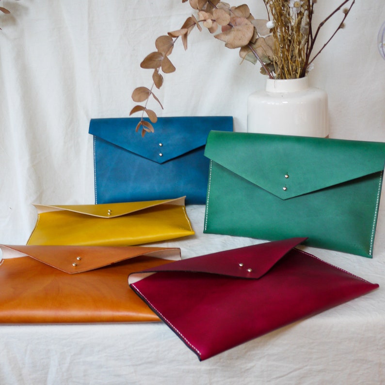 May include: Five leather envelopes in different colors: teal, green, yellow, brown, and burgundy. The envelopes are arranged on a white surface.