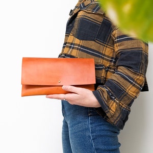 May include: A brown leather clutch with a flap closure and a gold button. The clutch is held by a person wearing a plaid shirt and blue jeans.