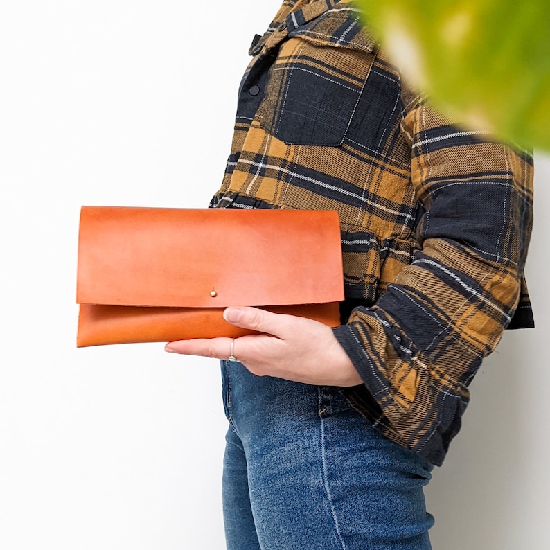 May include: A brown leather clutch with a flap closure and a gold button. The clutch is held by a person wearing a plaid shirt and blue jeans.