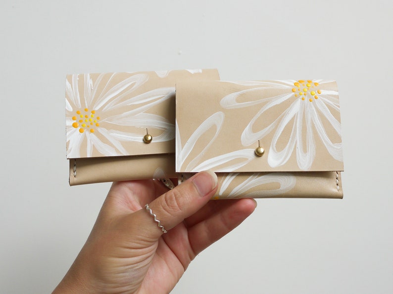May include: Two beige leather wallets with white daisy flower designs and yellow centers. The wallets have gold metal snaps.