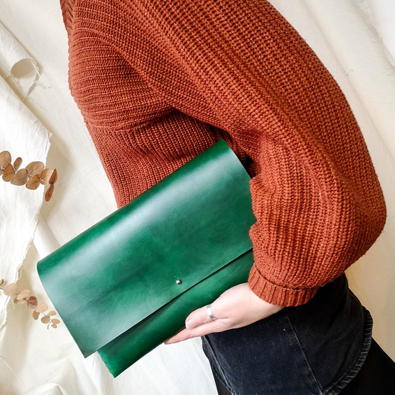 May include: A green leather clutch purse with a subtle flap closure. The purse is held by a person wearing a rust-colored sweater.