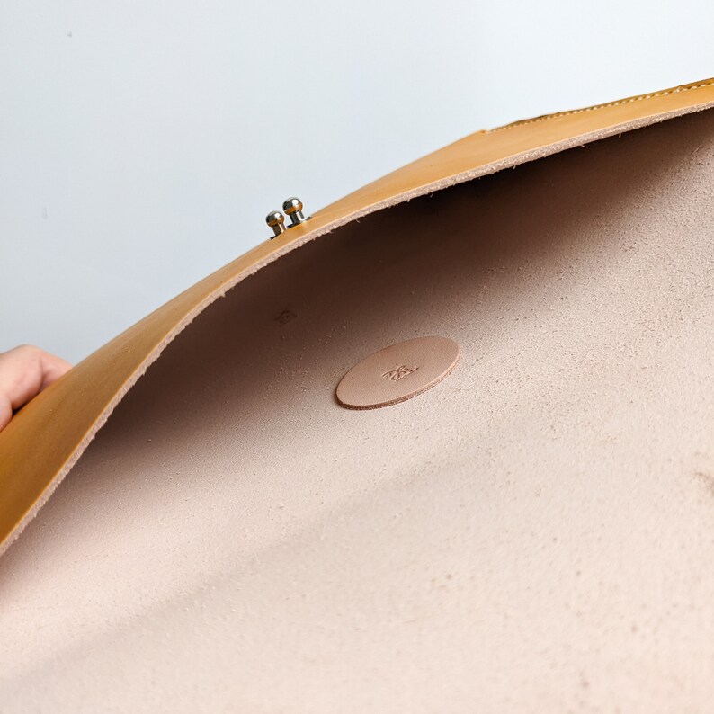 May include: A tan leather envelope with a silver clasp. The inside of the envelope is lined with a light tan fabric. The envelope has a small, round, tan leather label with the text "ZSL" embossed on it.