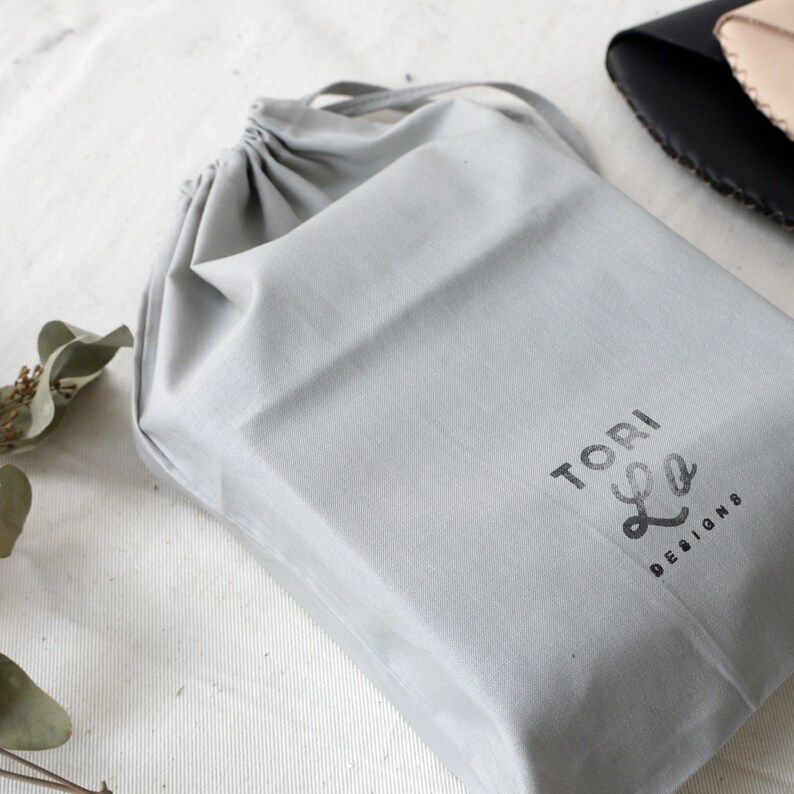 Op de afbeelding: Een grijze trekkoordtas met de tekst "TORI Lee DESIGNS" gedrukt op de voorkant.