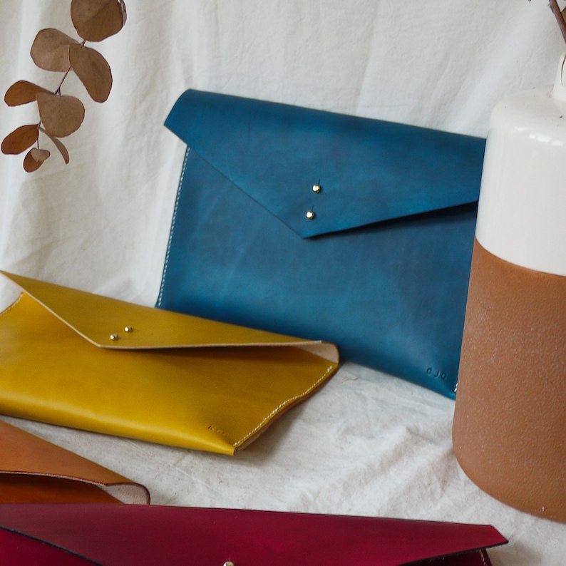 May include: Four leather envelope clutches in different colors: blue, yellow, brown, and red. The clutches have a flap closure and are made of a soft, supple leather. The clutches are arranged on a white fabric background.