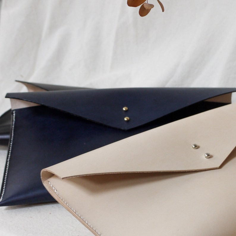 May include: Two leather envelopes, one in a light beige color and one in a dark blue color. Both envelopes have a flap closure and are secured with metal studs.