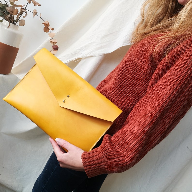 May include: A mustard yellow leather envelope clutch with a flap closure and two silver studs. The clutch is held by a person wearing a rust colored sweater.